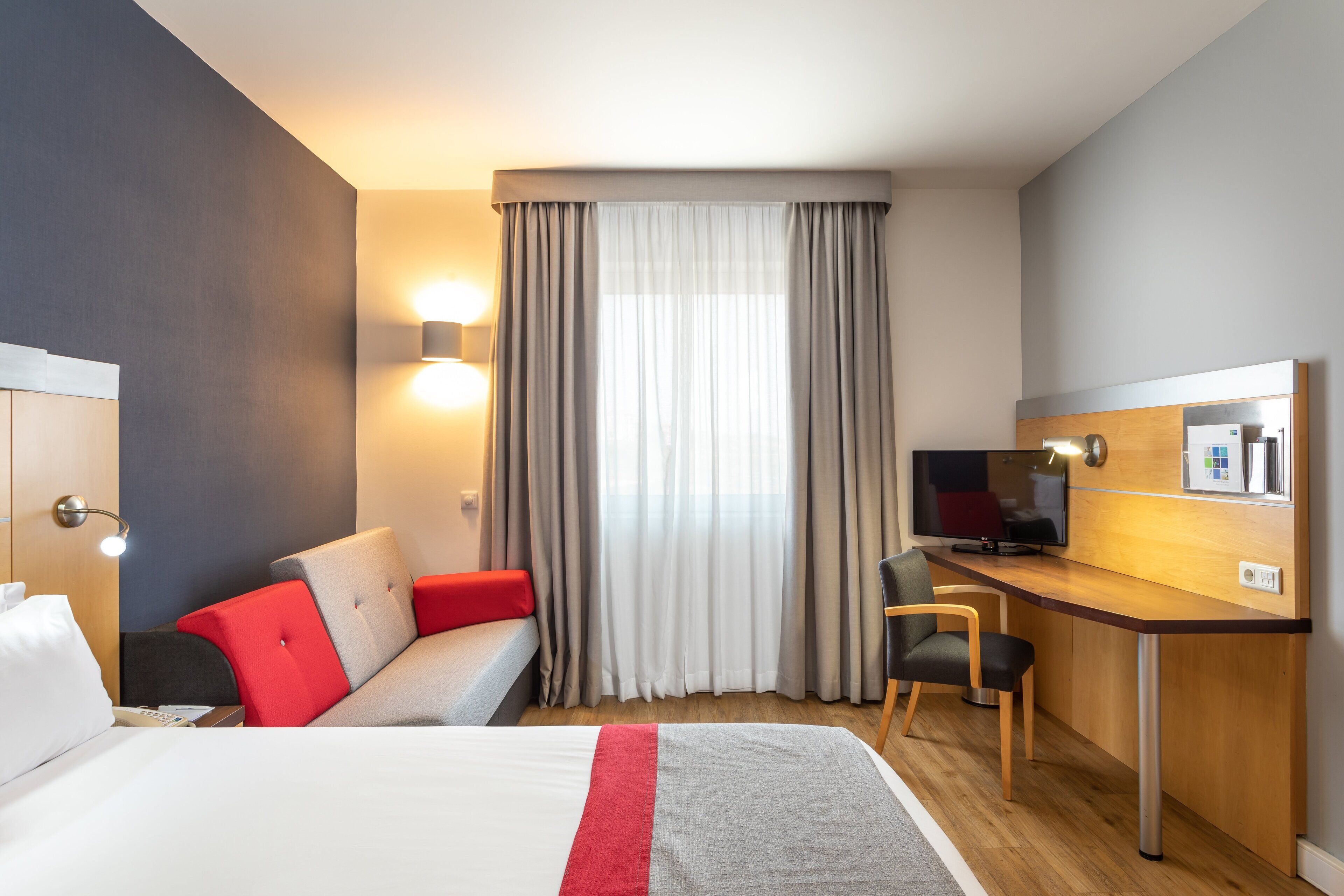 A hotel room with a bed, a desk with a computer, a chair, a sofa with red cushions, and a window with curtains.