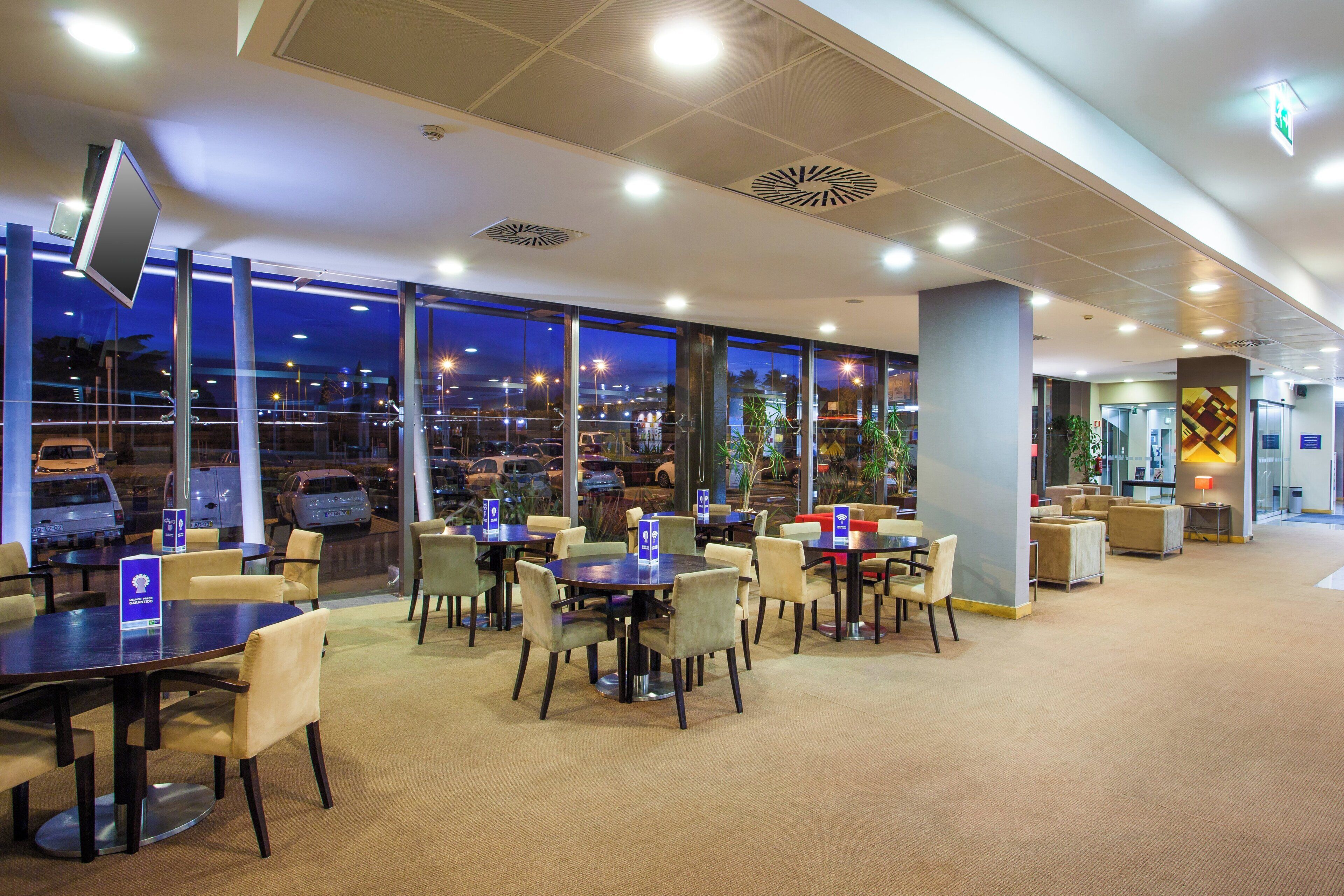 Un hall d’hôtel donnant sur le parking, avec plusieurs espaces aménagés avec des tables et des chaises, et un grand écran fixé au mur.