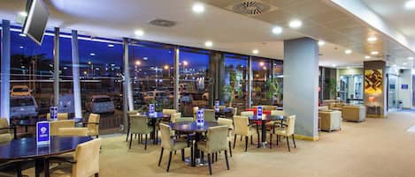 Un hall de hotel con vista a coches estacionados afuera, varias zonas de estar con mesas y sillas, y una pantalla grande montada en la pared.