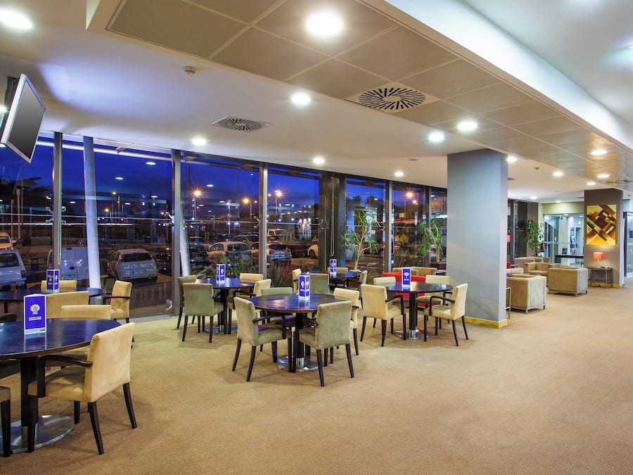 Un hall de hotel con vista a coches estacionados afuera, varias zonas de estar con mesas y sillas, y una pantalla grande montada en la pared.