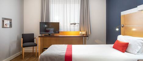 Habitació d'hotel amb un llit, escriptori amb televisor, cadira i finestra amb cortines.