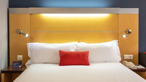 Habitación de hotel con cabecera de madera, ropa de cama blanca, almohada decorativa roja, teléfono en la mesita de noche y un cartel que indica el tipo de habitación.