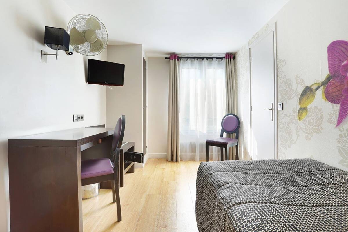 Een hotelkamer met een bed, een bureau met stoel, een televisie, een ventilator en een raam met gordijnen.