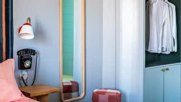 A hotel room with a bed, a wooden nightstand, a telephone, a mirror, a wardrobe, and a checkered ottoman.