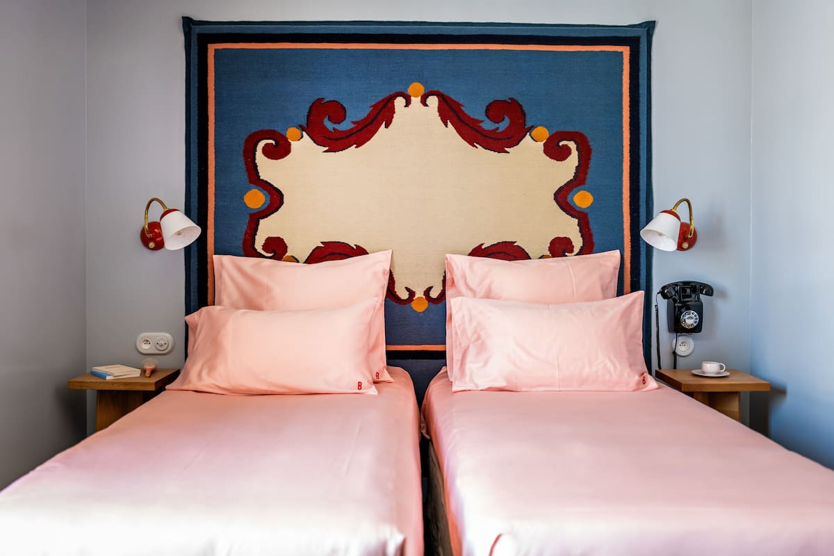 Een tweepersoonskamer met twee bedden, elk met roze beddengoed, een groot decoratief hoofdeinde, nachtkastjes met lampen en een vintage telefoon.