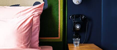 A neatly made bed with pink bedding, a small wooden nightstand with a vintage telephone and a glass of water, and a wall with a colorful abstract design.