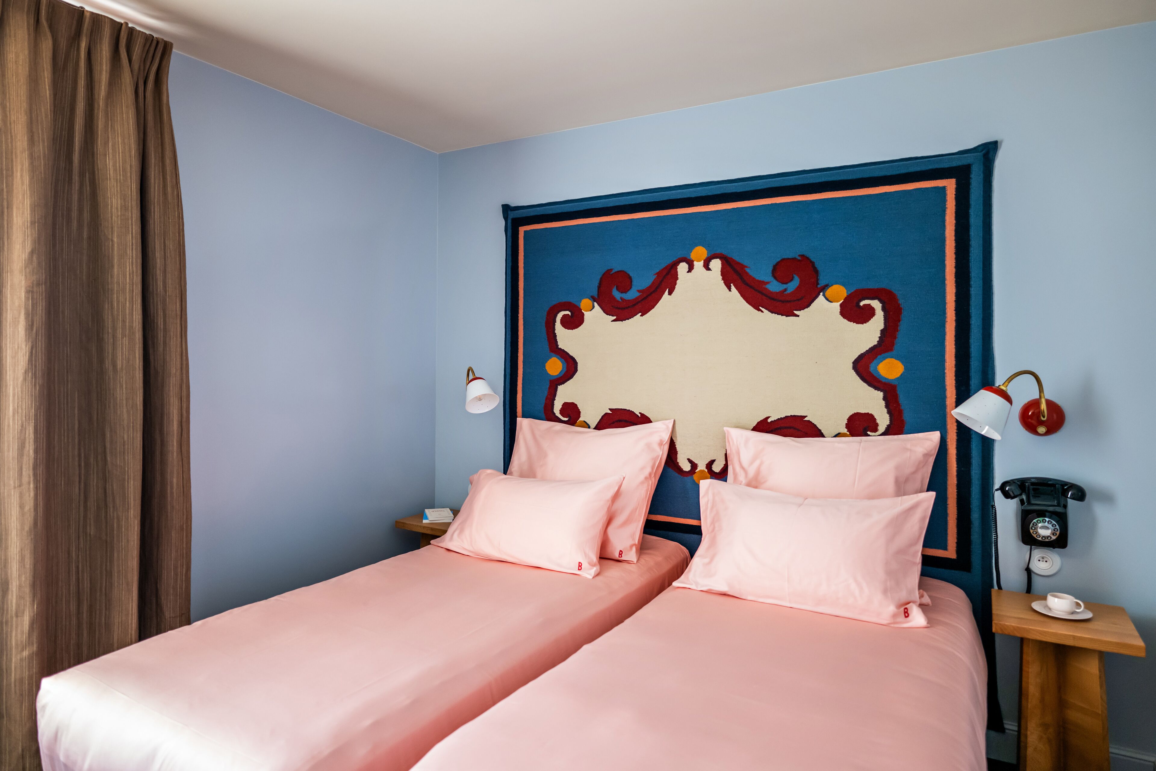 A double bed with pink bedding, a decorative headboard, bedside lamps, and a telephone on a wooden nightstand.
