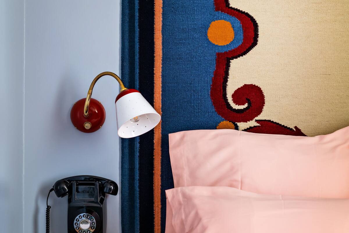 Een kamer met een antieke draaischijf telefoon, een witte lamp, een bed met roze beddengoed en een kleurrijke wandtapijt.
