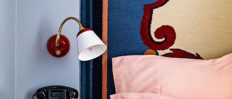 A room with a vintage rotary phone, a white lamp, a bed with pink bedding, and a colorful tapestry on the wall.