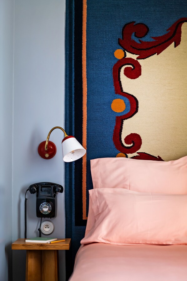 Une pièce avec un vieux téléphone à cadran, une lampe blanche, un lit avec une literie rose et une tapisserie colorée au mur.