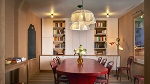 Un comedor con una mesa roja, sillas, un jarrón con flores y estanterías.