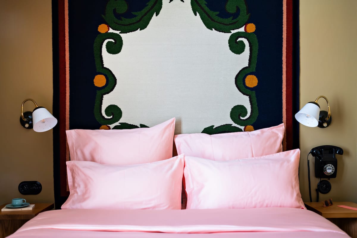 Een bed met roze beddengoed, een decoratief wandtapijt en een telefoon in vintage-stijl op een nachtkastje.
