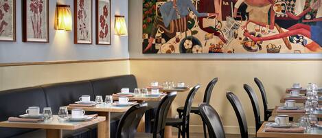A restaurant with wooden tables and black chairs, framed floral artwork on the wall, and a mural depicting people dining.