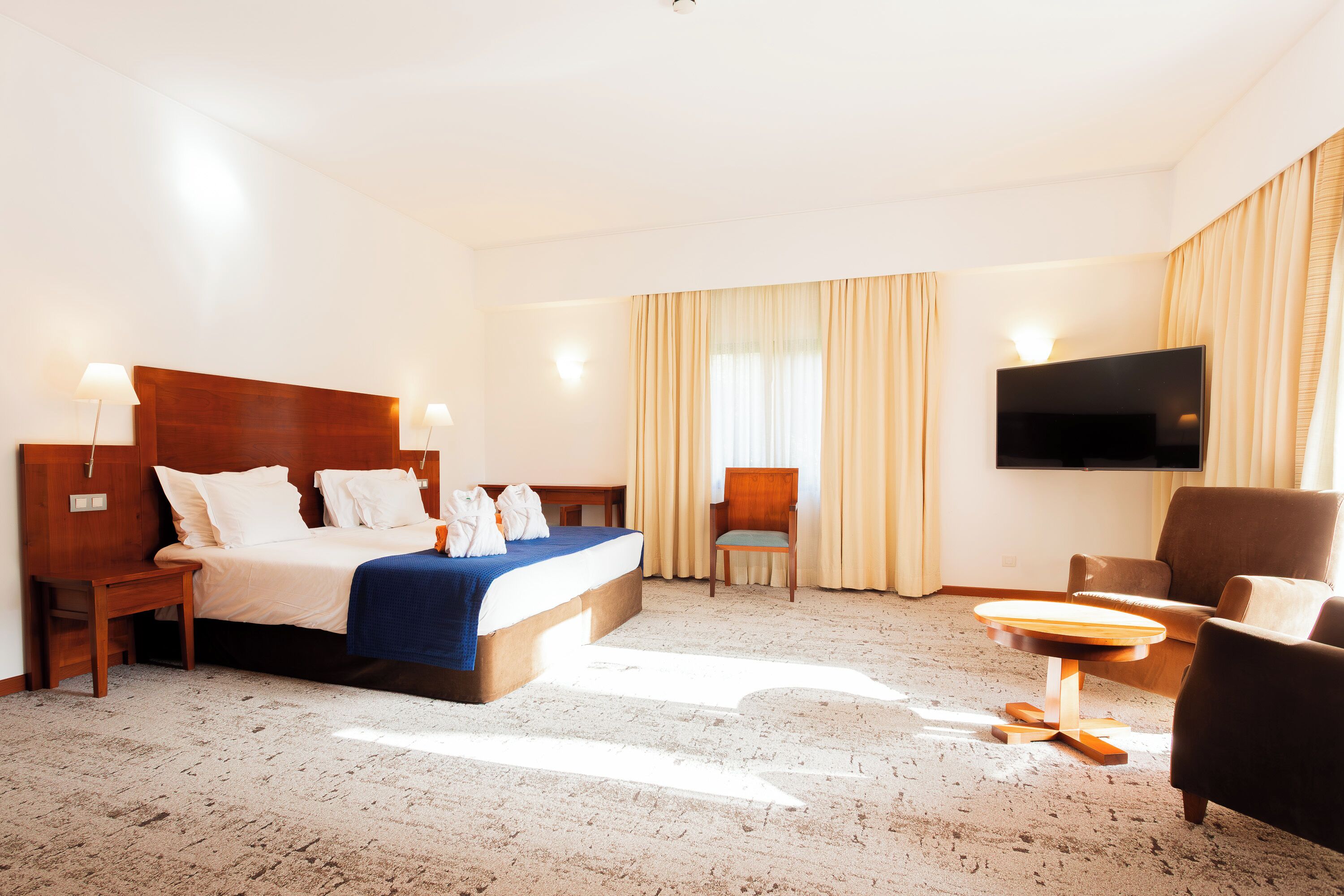 Une chambre d’hôtel avec un grand lit, un téléviseur fixé au mur, un coin salon avec une table et une fenêtre avec des rideaux.