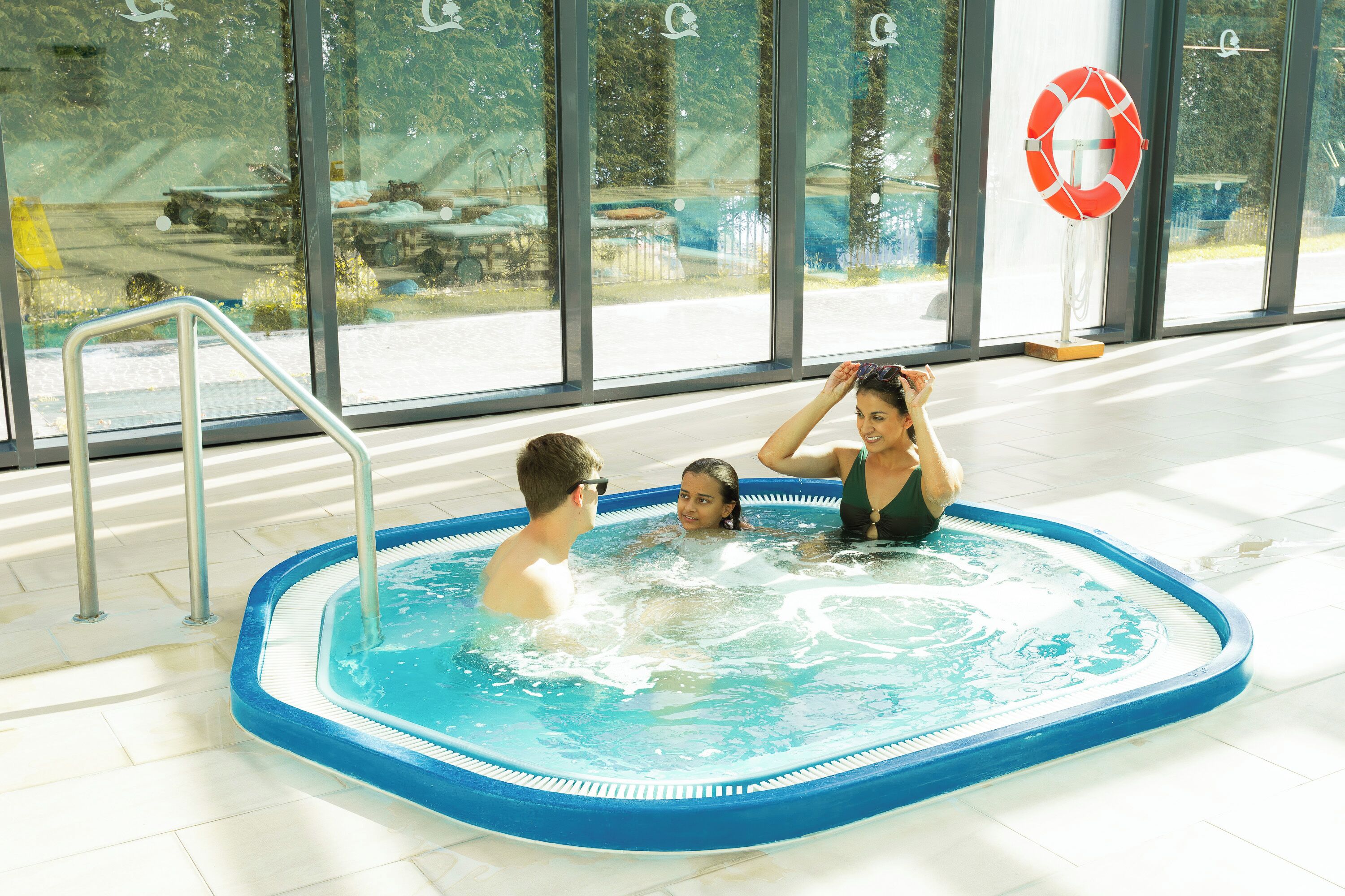 A swimming pool with a lifebuoy, metal ladder, and a view of the outside through large windows.