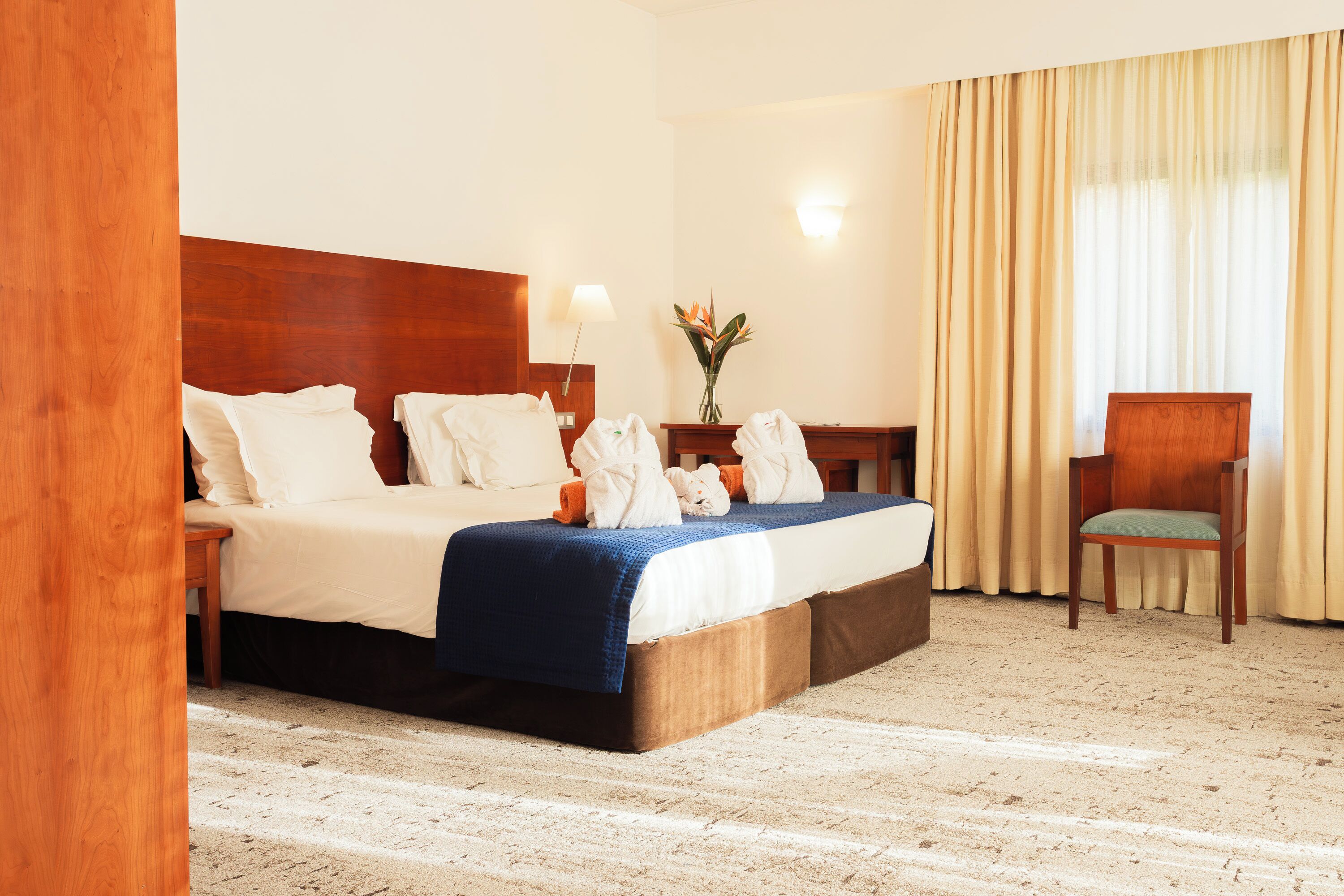 A hotel room with a large bed, a wooden headboard, a nightstand, a desk, a chair, and a vase with flowers.