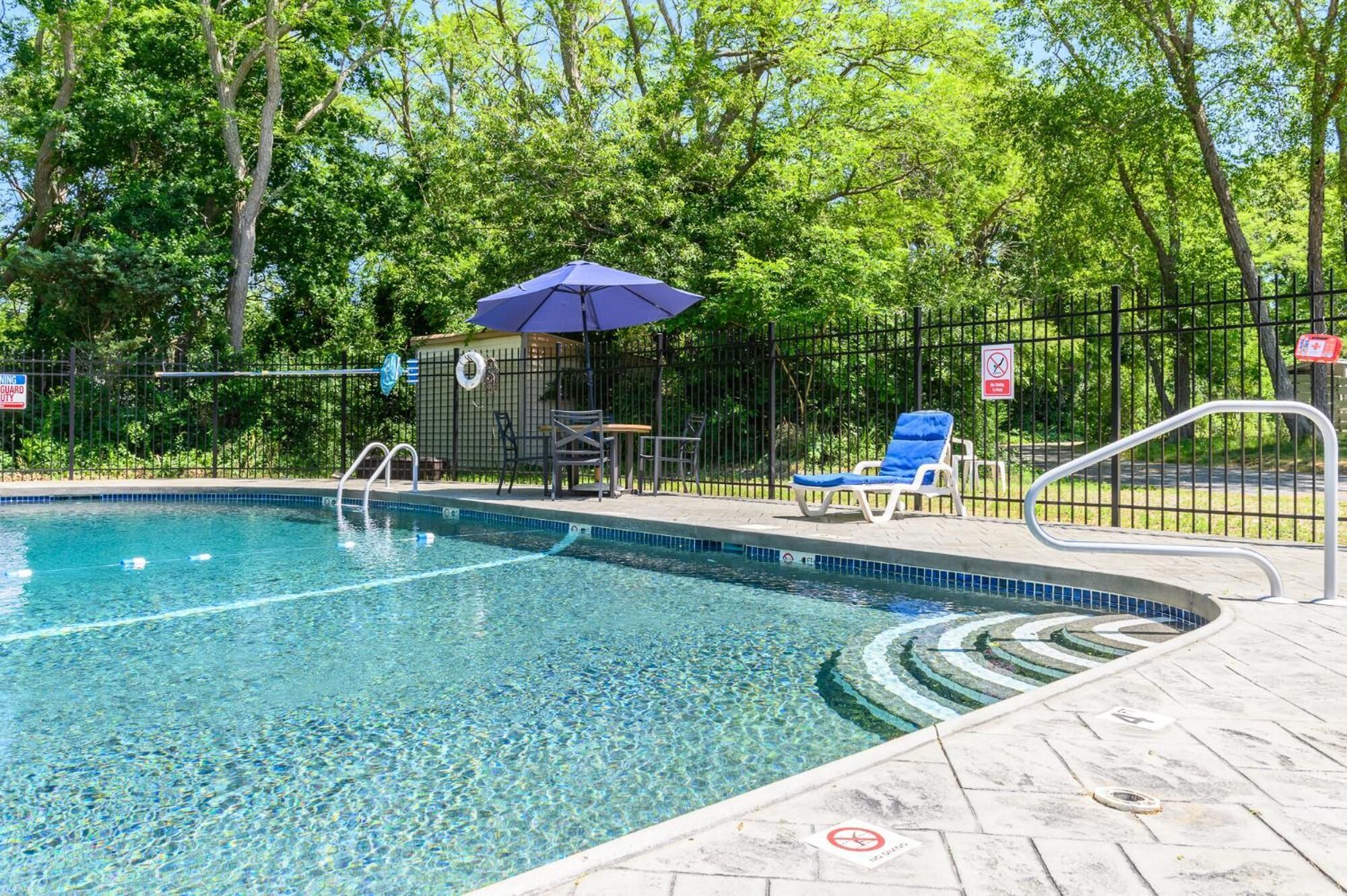 A clear swimming pool with a blue umbrella, a lifeguard chair, and a fenced area with a no swimming sign.