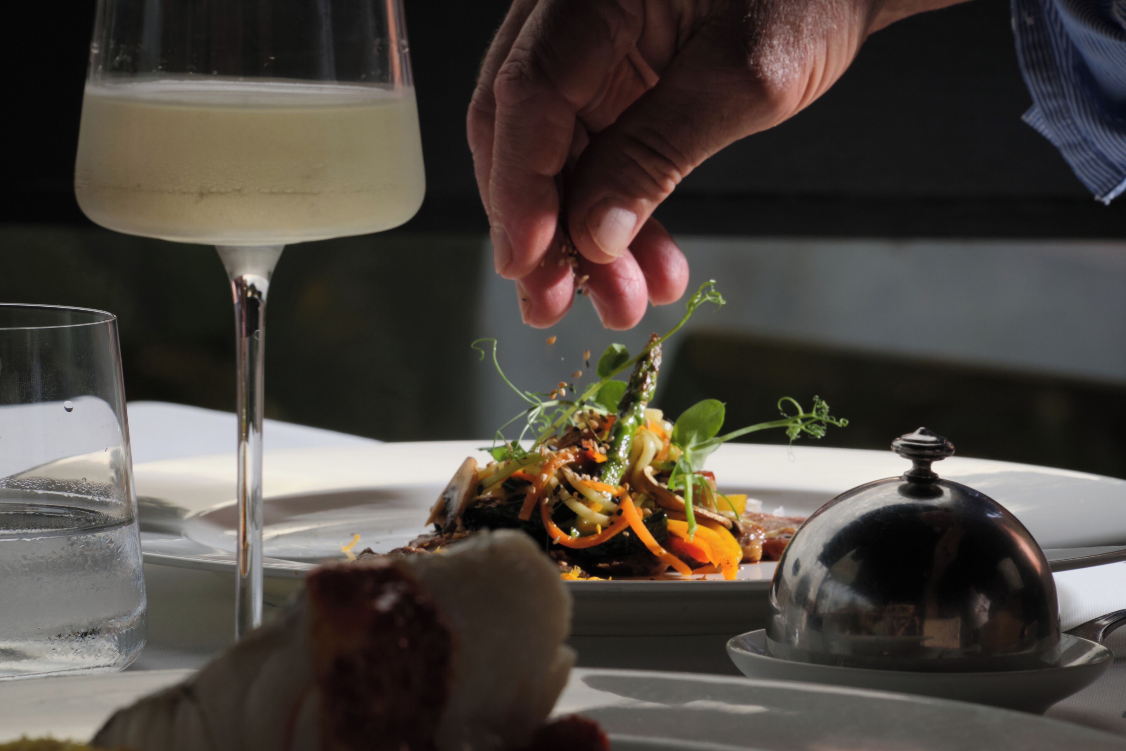 A dining table with a glass of white wine, a plate of food, and a hand sprinkling seasoning.