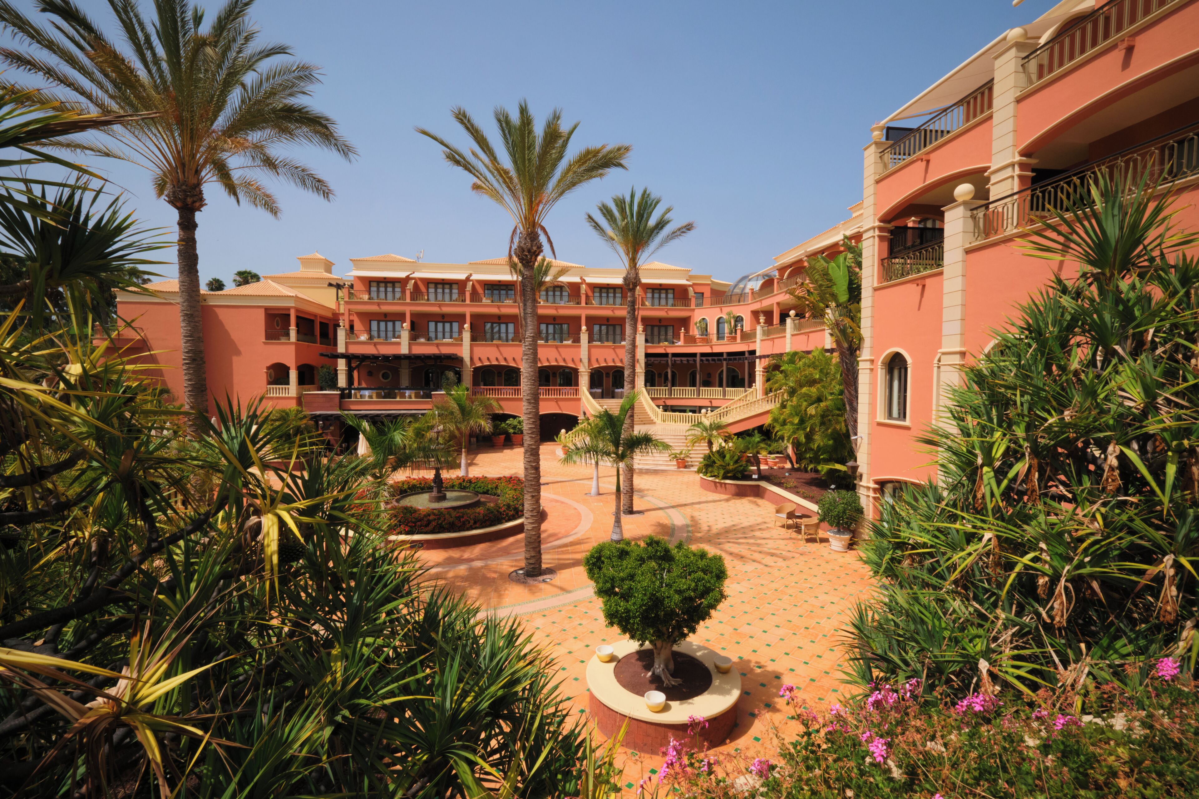 A hotel courtyard with palm trees, a central fountain, and a tiled floor.