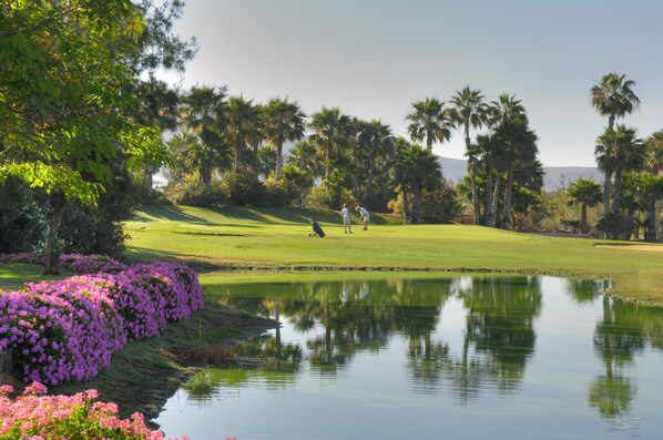 Un campo de golf con un estanque, palmeras y flores moradas.