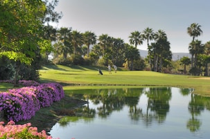 Un campo de golf con un estanque, palmeras y flores moradas.