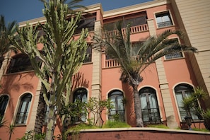 A multi-story building with balconies, windows with bars, and various plants including cacti and palm trees.