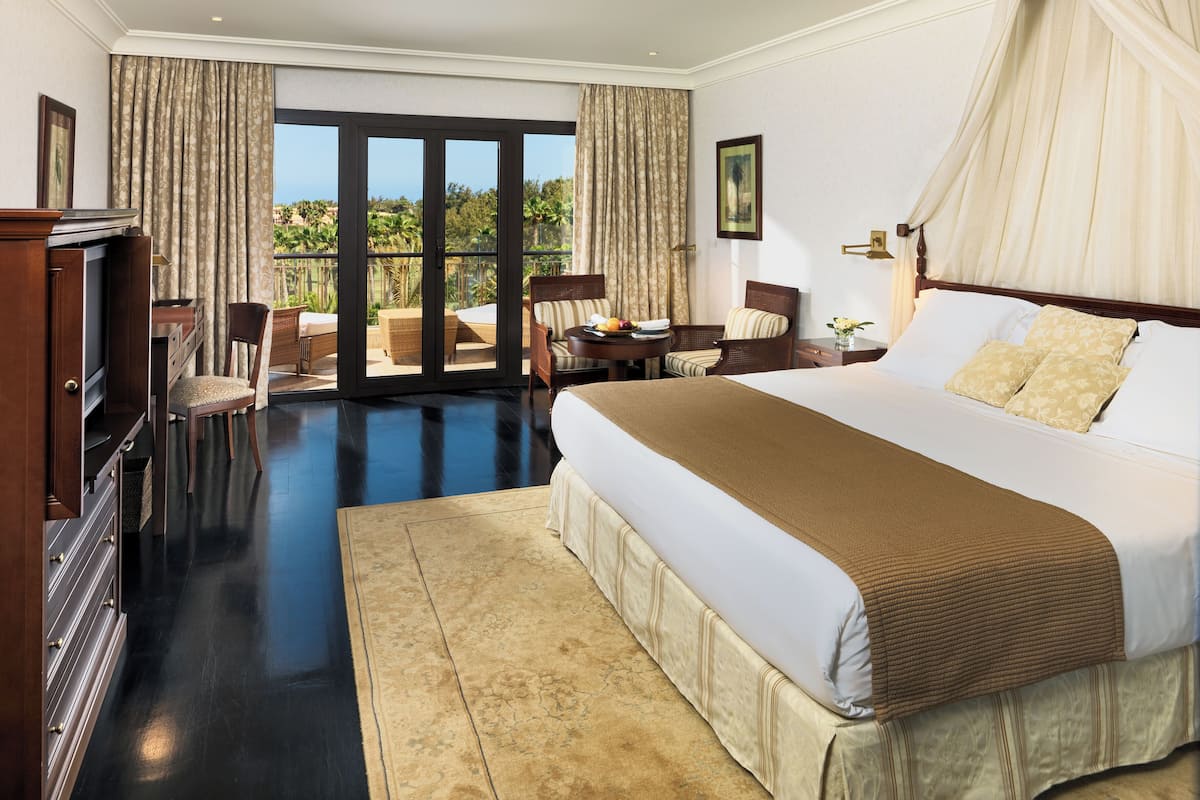 Ein Hotelzimmer mit einem großen Bett, einem Fernseher, einem Schreibtisch mit Stuhl und einem Balkon mit Gartenmöbeln.