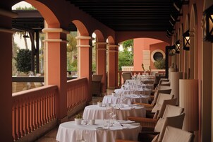 Un restaurante con mesas y sillas redondas, cubierto por un techo con arcos, y vistas al exterior con vegetación.