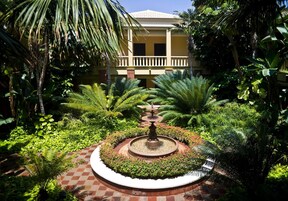 Un patio con una fuente central, rodeado de abundante vegetación y un edificio con balcón.