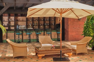 Una zona de bar con mobiliario de mimbre, un paraguas grande y piso de baldosas.