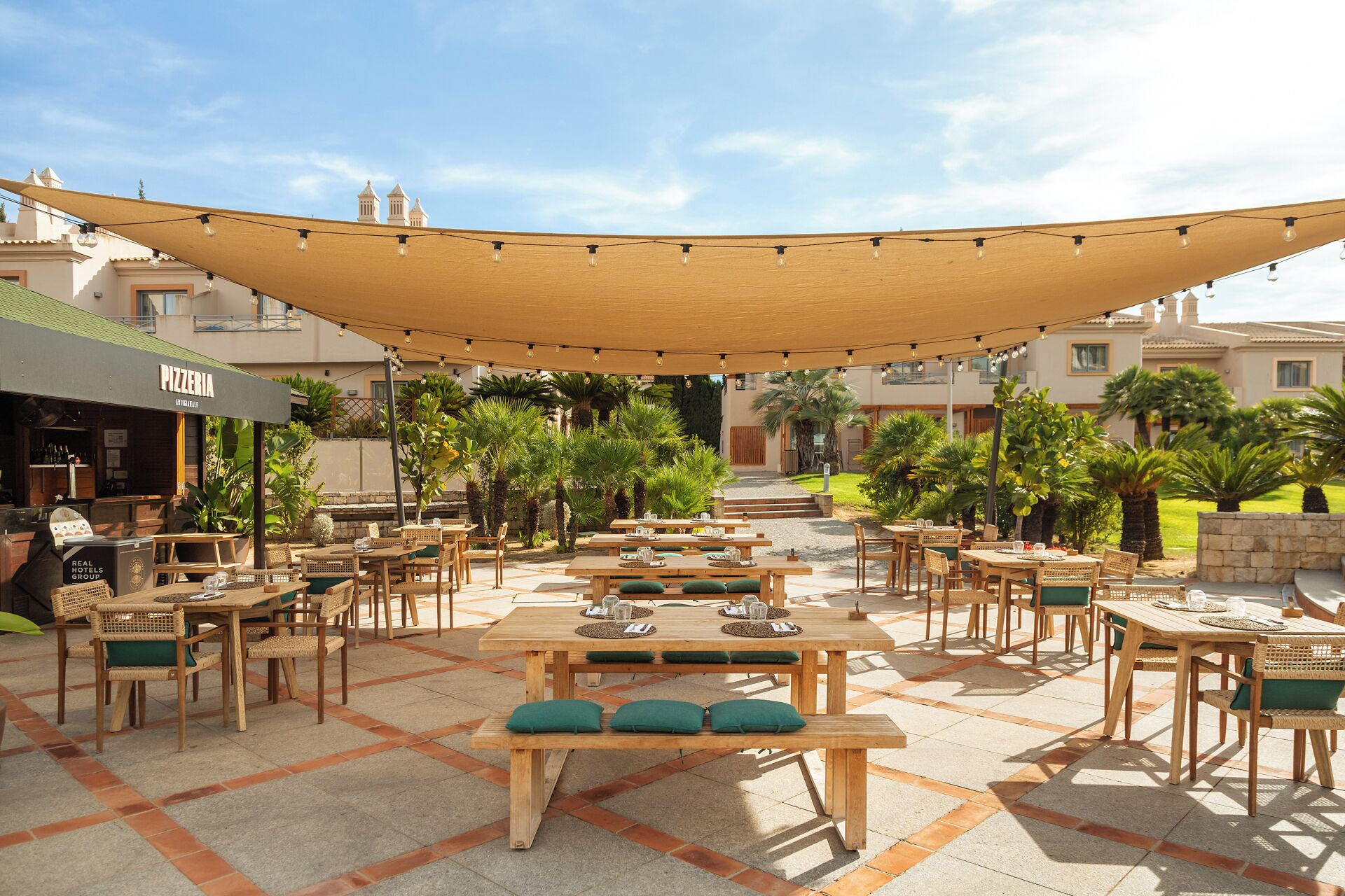 Un espacio al aire libre con varias mesas y sillas de madera, un toldo sombreado y una pizzería.