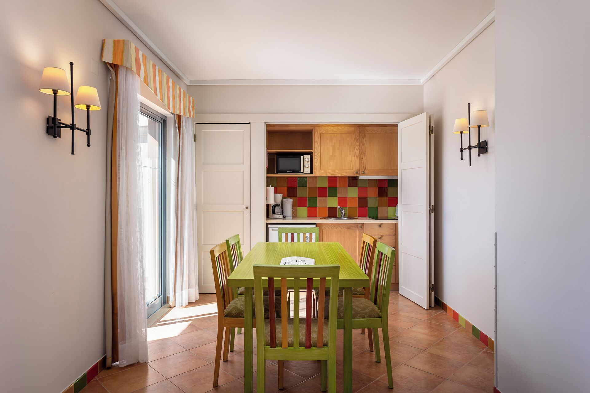 A dining area with a green table and chairs, a kitchen with colorful tiles, and a microwave.