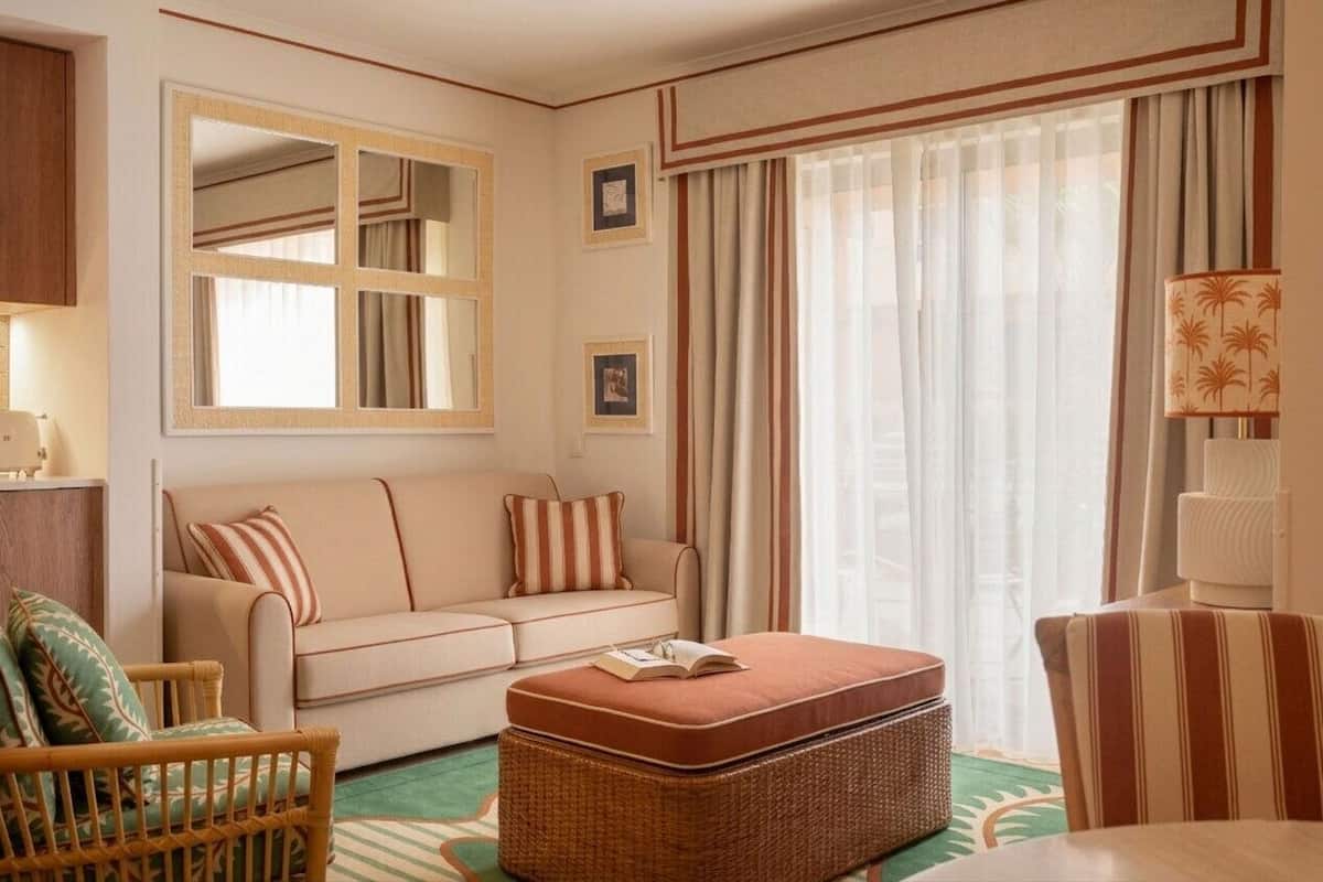 A hotel room with a sofa, ottoman, wicker chair, and a lamp.