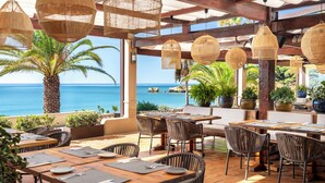 A restaurant with outdoor seating overlooking the sea, featuring wicker chairs and tables, and hanging wicker lamps.