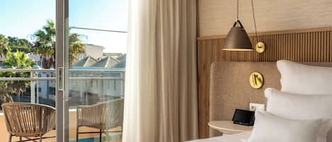 Habitación de hotel con cama, mesita de noche con lámpara, una silla y vistas al balcón exterior.
