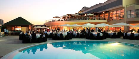 A poolside dining area with tables and umbrellas, a covered seating area, and a large building in the background.