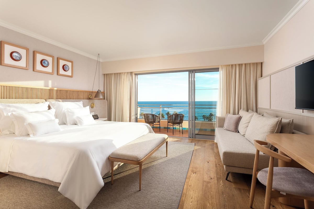 A hotel room with a large bed, a sofa, a desk, and a balcony with a view of the ocean.