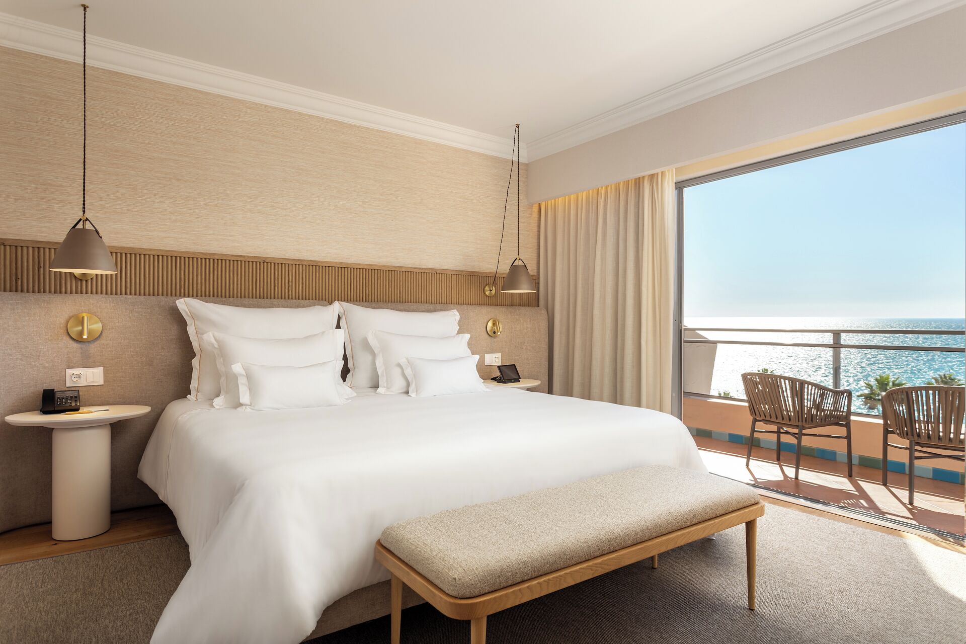 A hotel room with a large bed, bedside tables, a bench, and a balcony with a view of the sea.