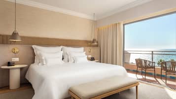 A hotel room with a large bed, bedside tables, a bench, and a balcony with a view of the sea.