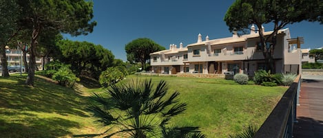A modern hotel with a well-maintained lawn, large trees, and a clear blue sky.