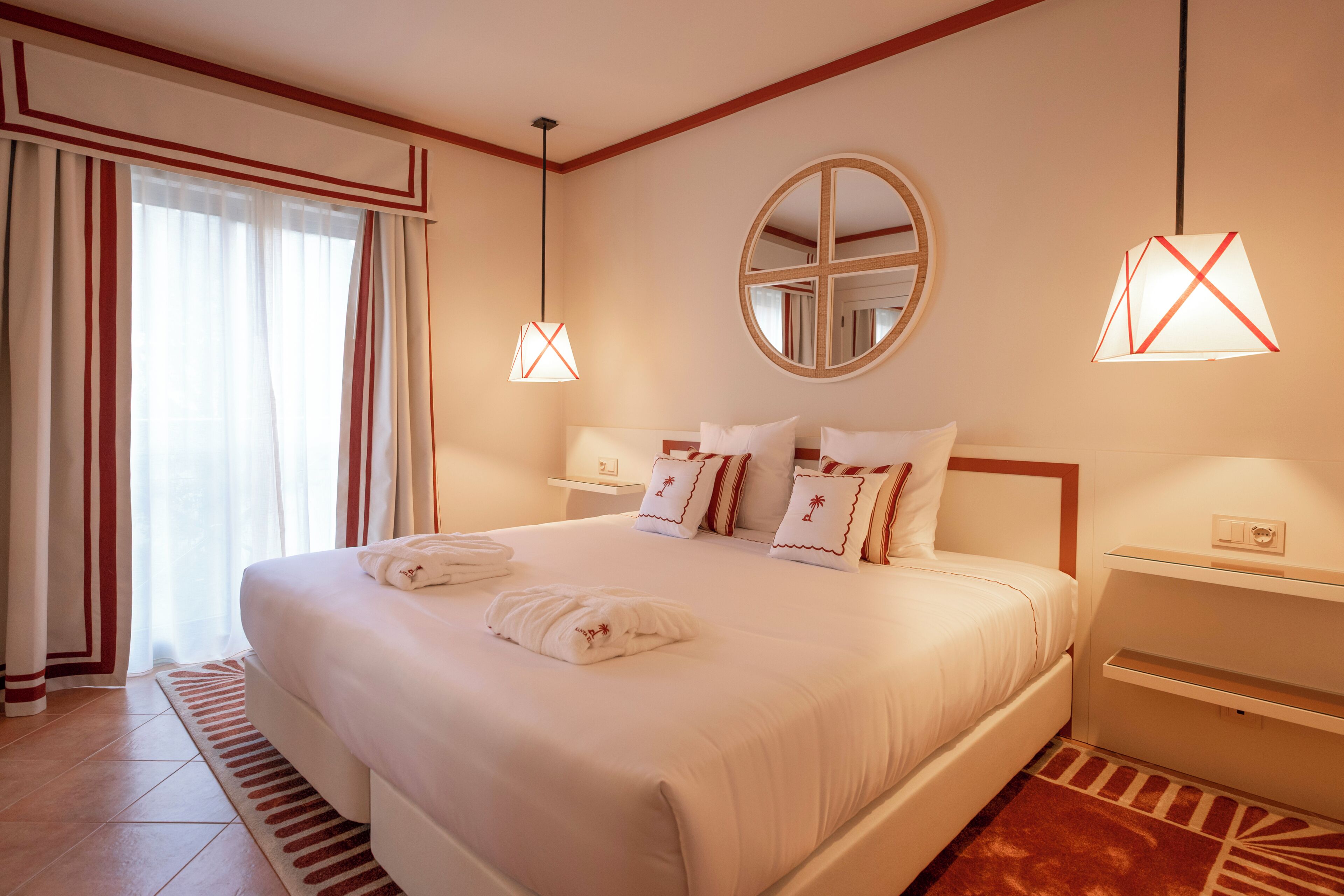 Une chambre d’hôtel avec un grand lit, deux tables de chevet, un miroir rond et une fenêtre avec des rideaux.