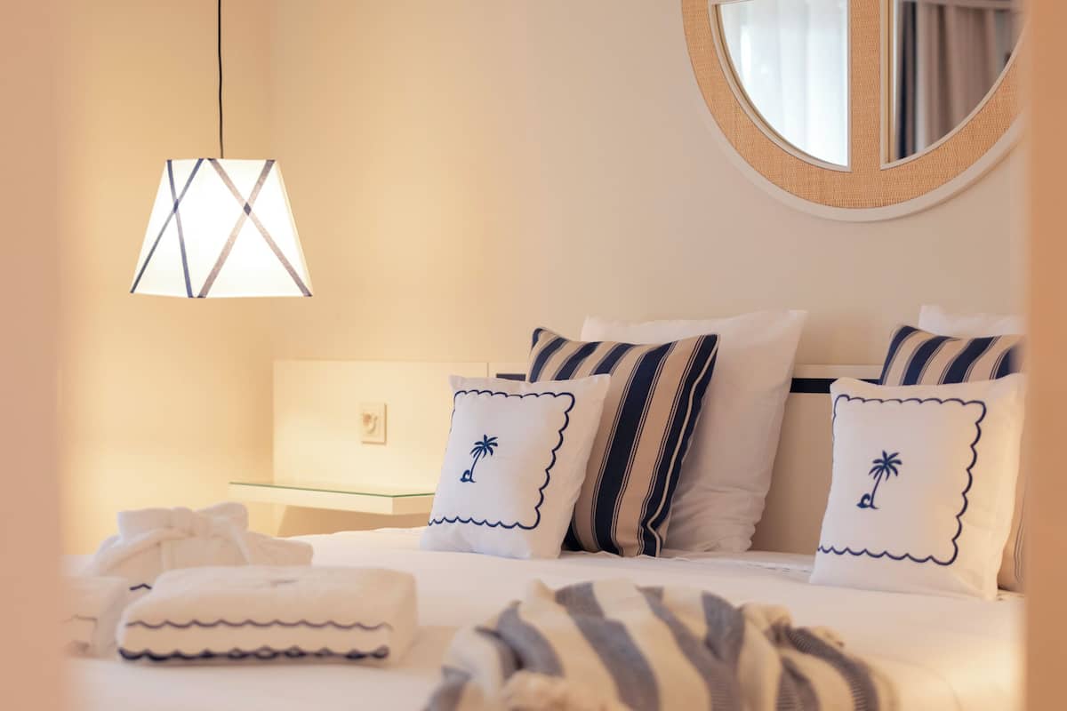 A neatly arranged bedroom with a bed, pillows, a bedside table, a lamp, and a circular mirror on the wall.
