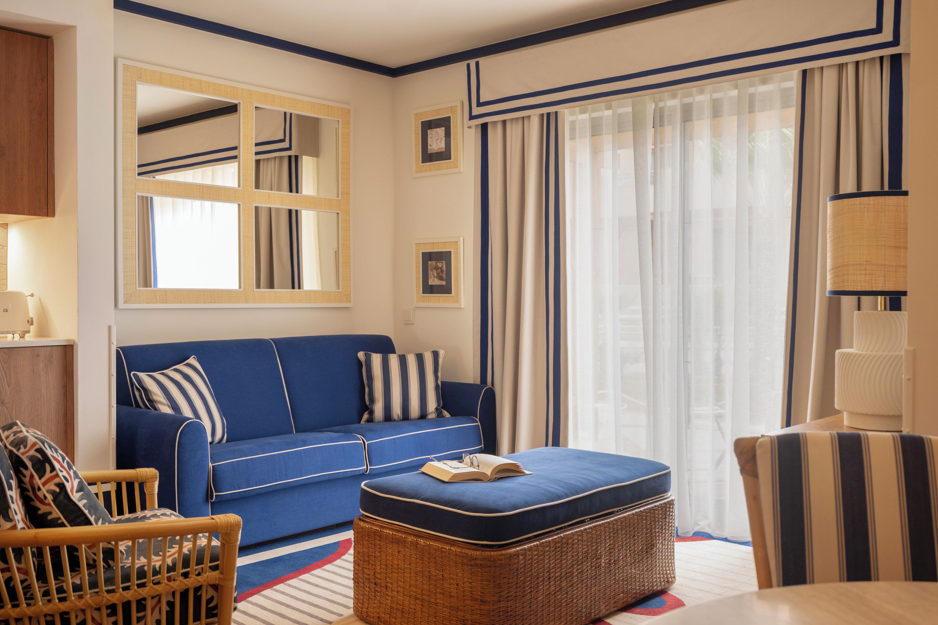 A room with a blue sofa, a wicker ottoman, a striped chair, and a lamp.