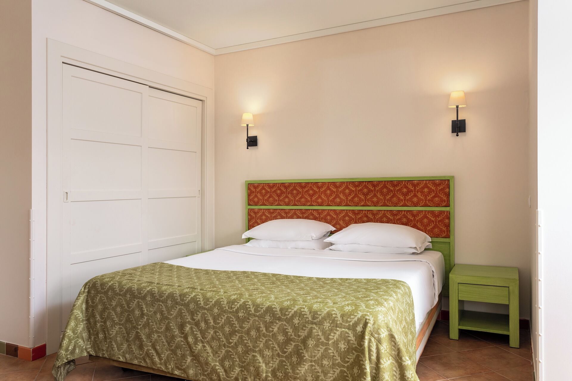 A bedroom with a bed, a green nightstand, two wall-mounted lights, and a white door.
