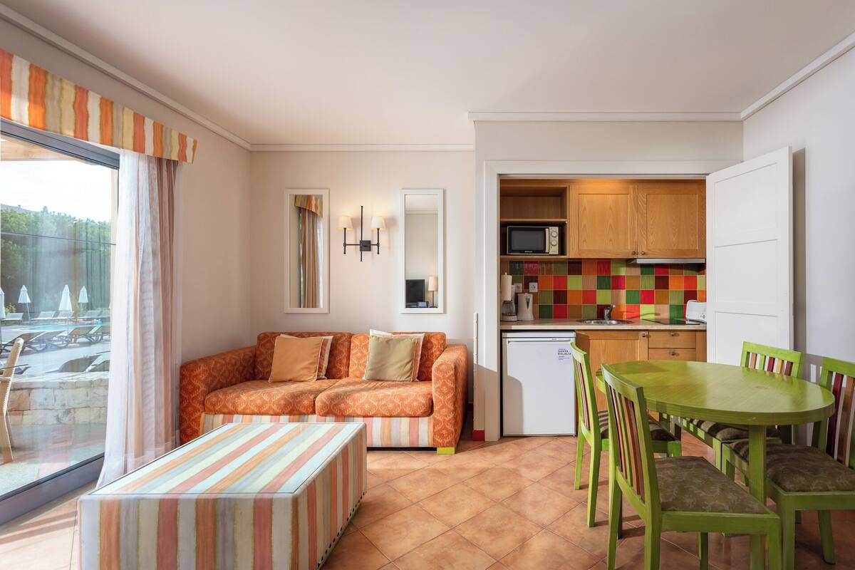 A compact living space with a sofa, a dining table with chairs, a kitchenette, and a view of an outdoor swimming area.