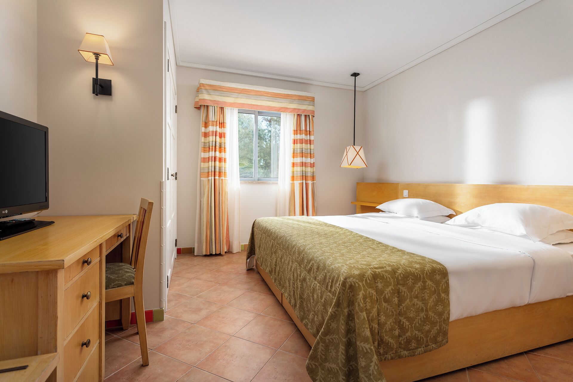 Habitación de hotel con cama, escritorio con computadora, silla, ventana con cortinas y lámpara colgante.