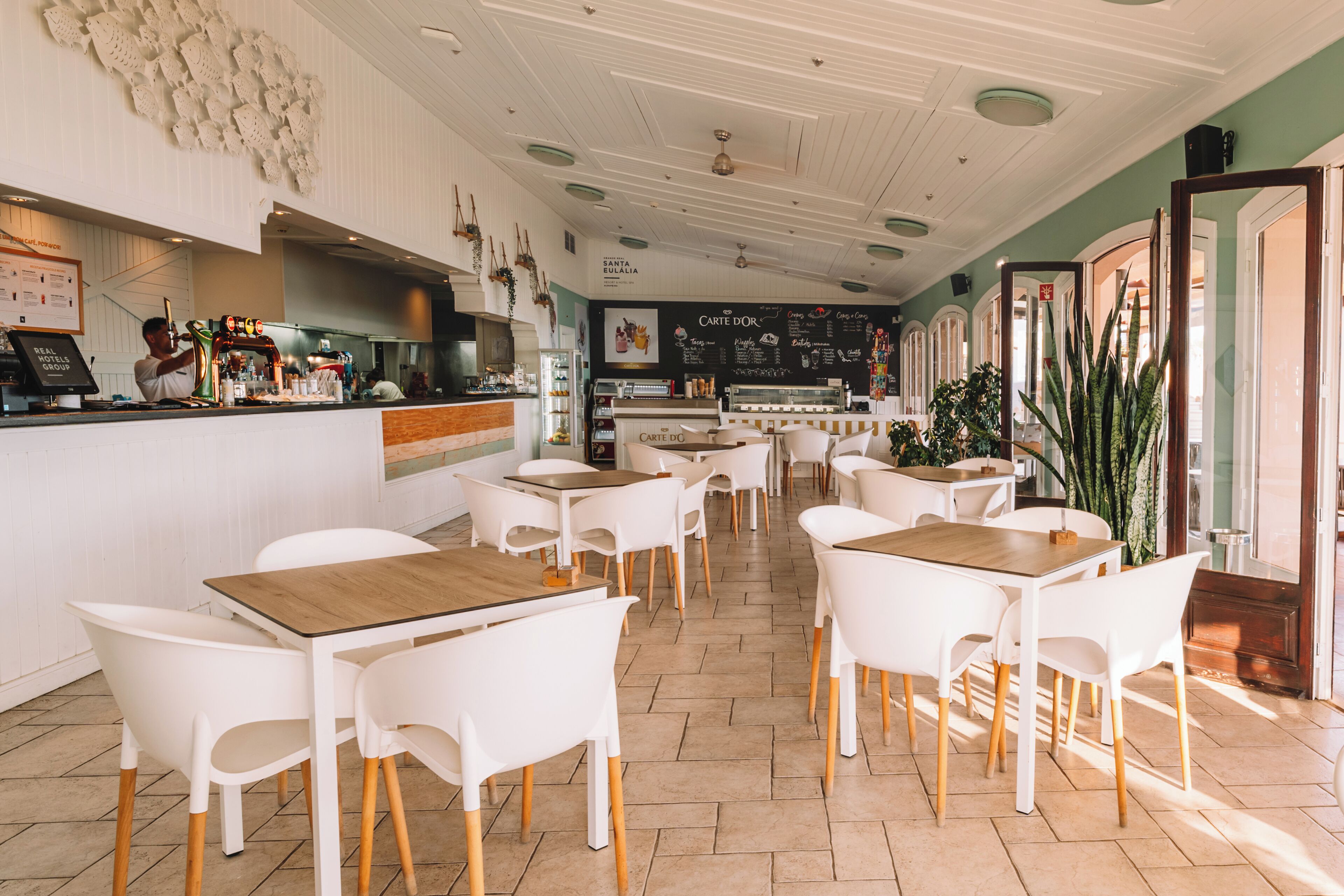 A restaurant interior with white chairs and tables, a bar area, and a chalkboard menu.