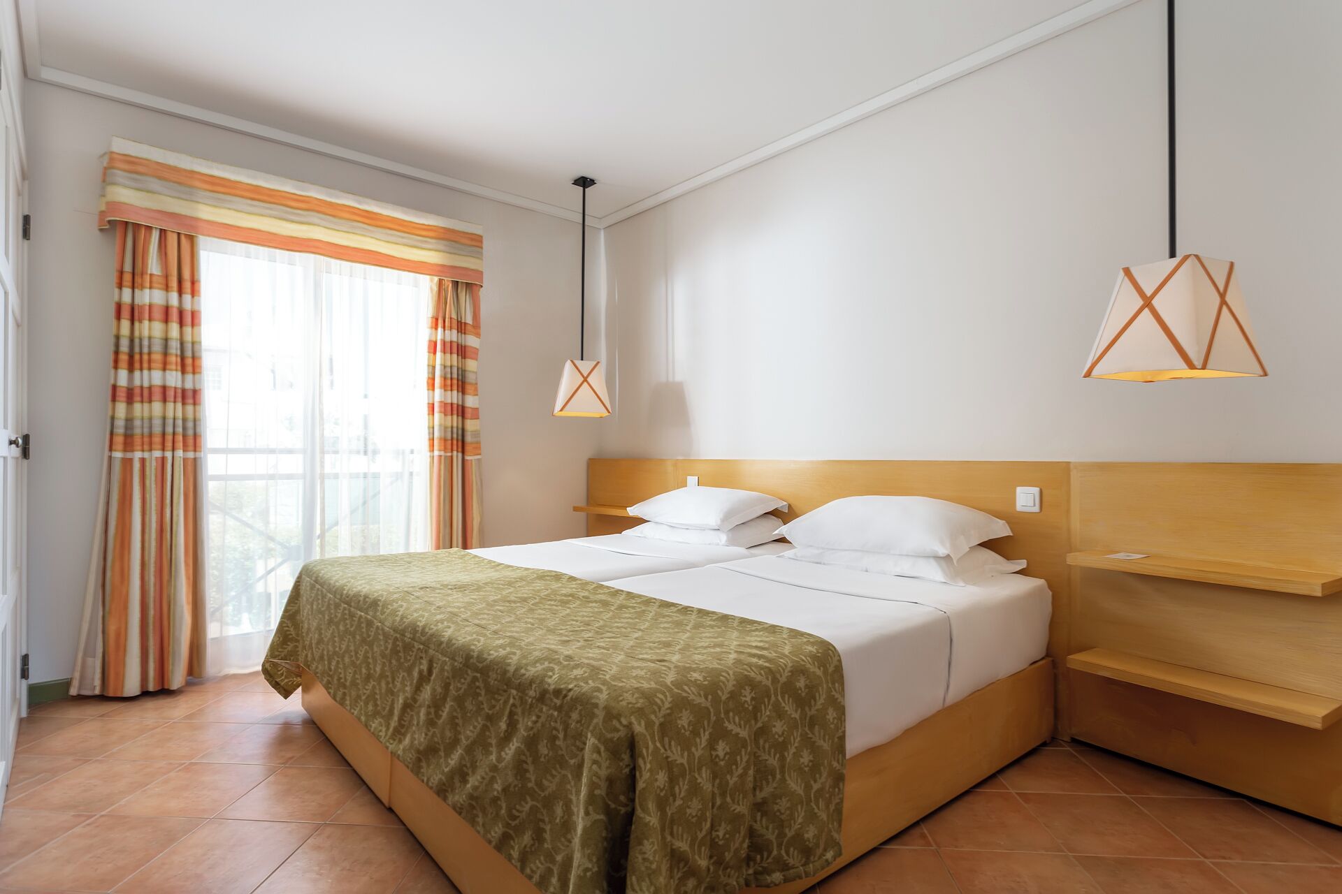 Een hotelkamer met een tweepersoonsbed, een houten hoofdbord, een nachtkastje, een raam met gordijnen en een hangende hanglamp.