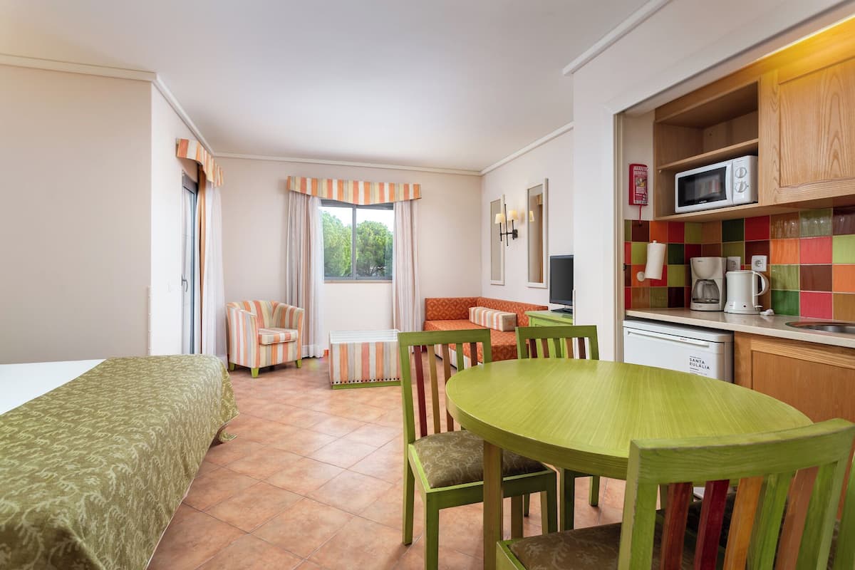 A compact room with a bed, a dining table with chairs, a kitchenette, and a window with curtains.