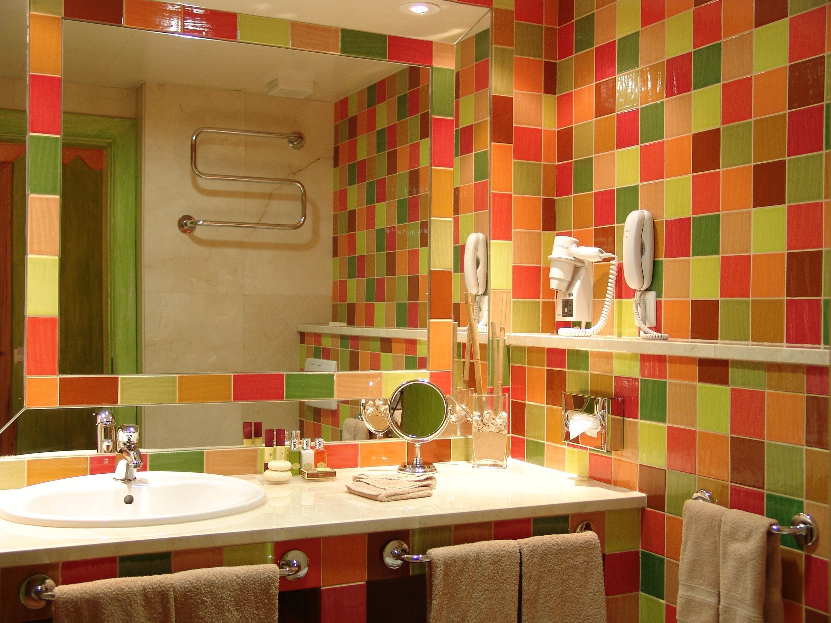 A bathroom with a colorful mosaic tile wall, a sink with a mirror, and a hairdryer on a shelf.
